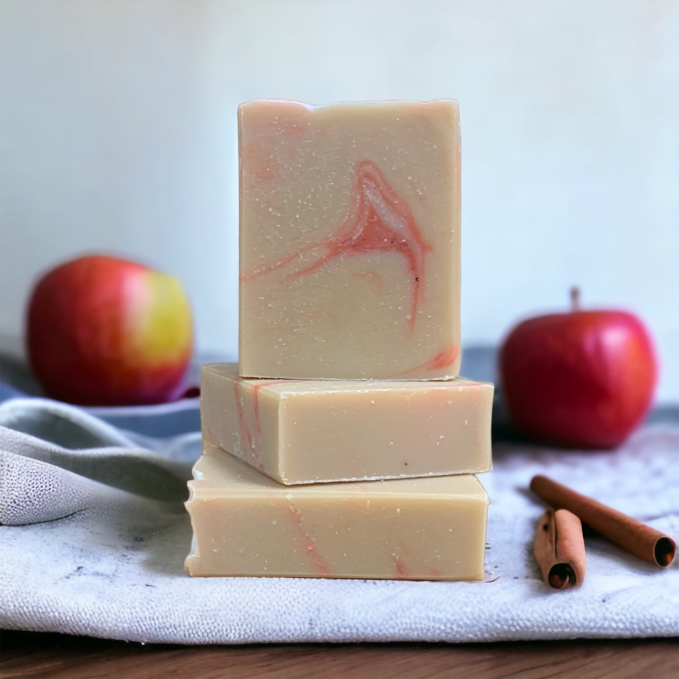A brown and red rectangular bar of soap on a wood countertop with apples, cinnamon sticks, and a towel in the background.