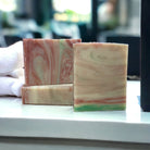 A white, red, and green rectangular bar of soap on a white countertop with a towel and a window in the background.